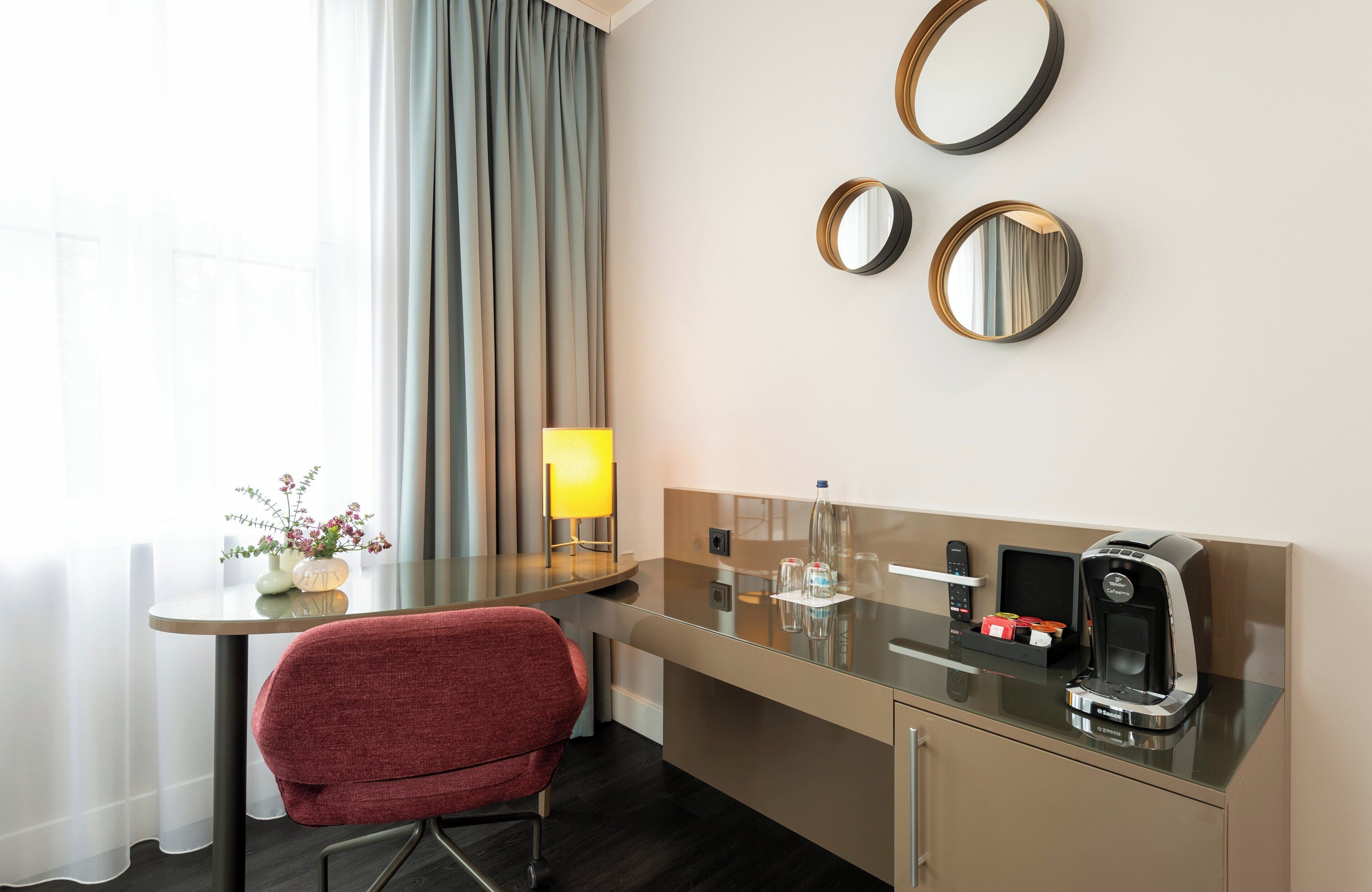 A hotel room with a desk, a chair, a coffee machine, and two wall-mounted mirrors.