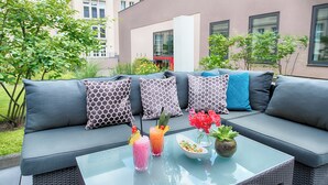 A modern outdoor seating area with a grey sofa, patterned cushions, and a glass table with drinks and a flower vase. - Leonardo Royal Hotel Berlin Alexanderplatz (Berlin)