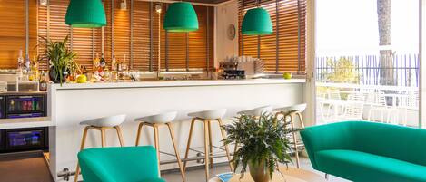 Zona de bar moderna con encimera blanca, luces colgantes verdes y persianas de madera. Hay taburetes de bar, una planta en maceta y vistas al balcón exterior.