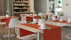 A modern dining area with white tables and chairs, orange tablecloths, and a ceiling with circular light fixtures.