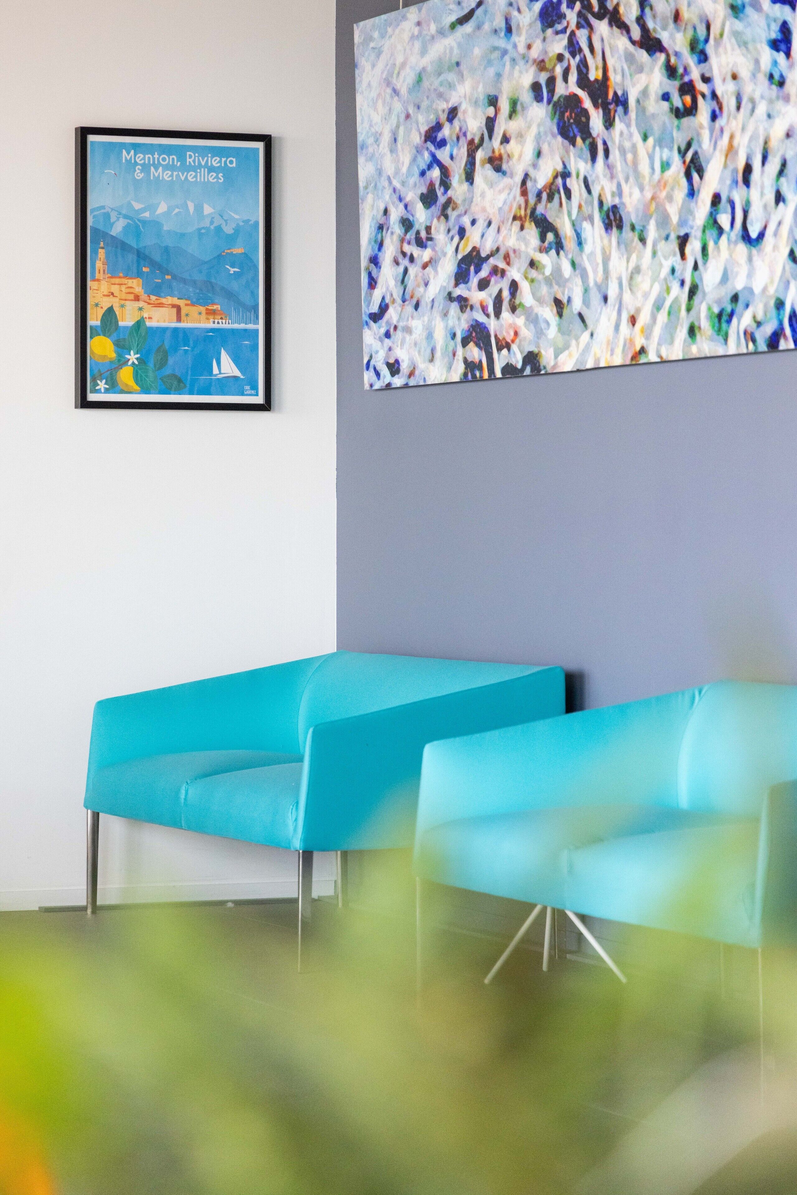 A room with two teal chairs, a framed picture of Menton, Riviera & Merveilles, and a large abstract painting.
