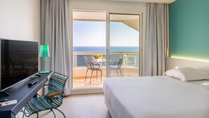 A hotel room with a bed, a desk with a TV and lamp, a chair, and a balcony with a view of the sea.