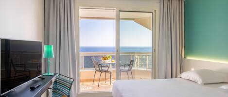 Habitación de hotel con cama, escritorio con televisor y lámpara, silla y balcón con vista al mar.
