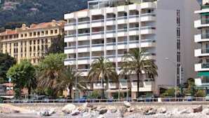 Un hotel frente al mar con varios pisos, una playa en primer plano y una montaña al fondo.