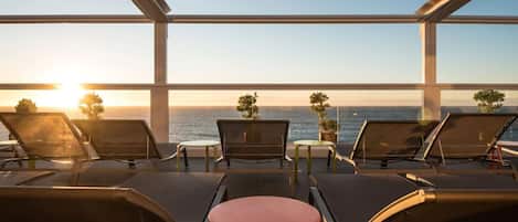 A sunlit terrace with lounge chairs and a small table, overlooking a body of water and a clear sky.