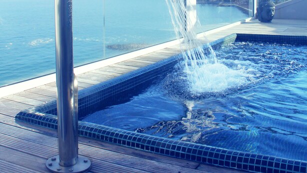 Un jacuzzi con grifo metálico, rodeado por barandilla de vidrio, con vista a una masa de agua.