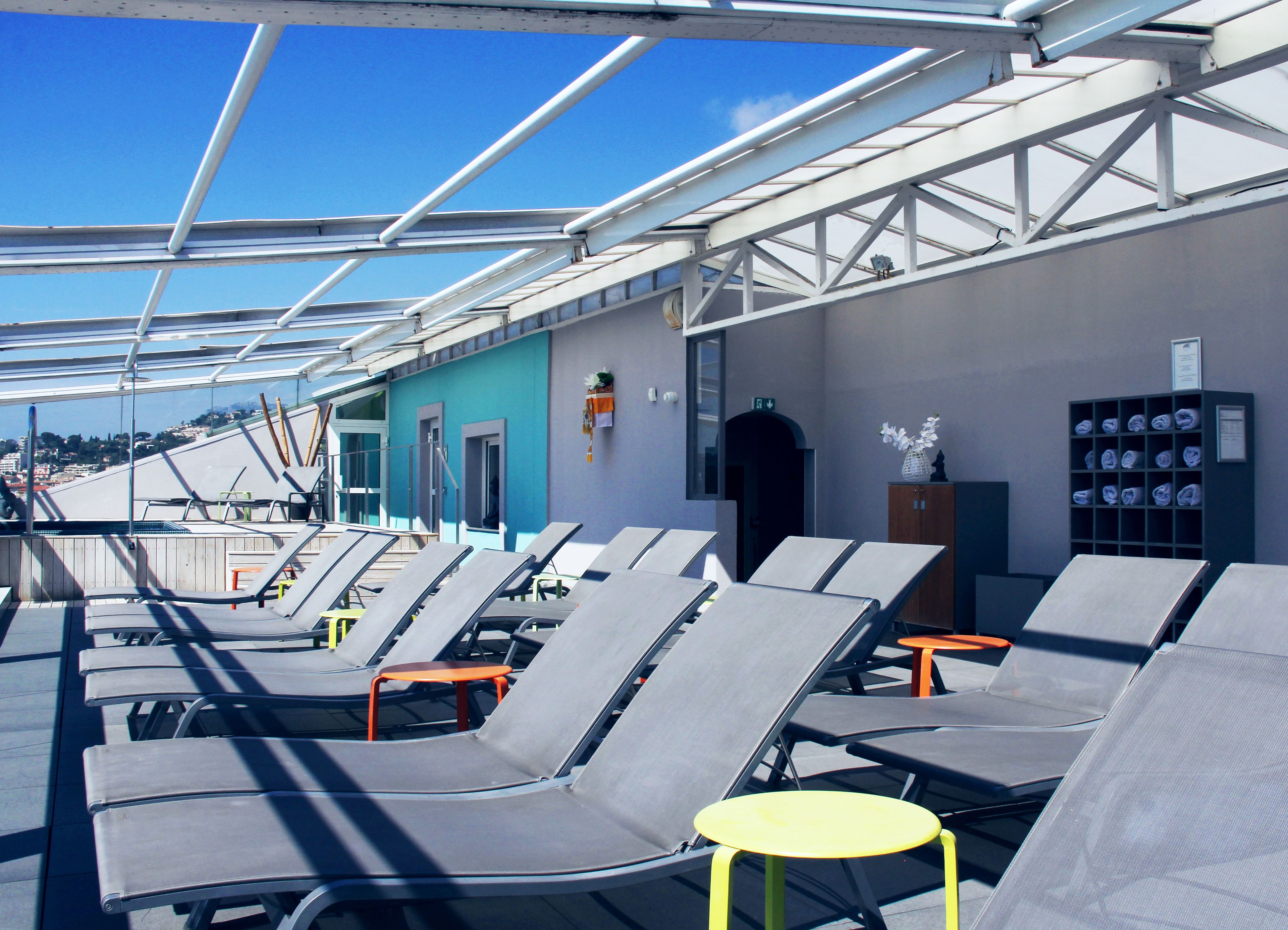 Una terrazza prendisole con lettini e un piccolo tavolo giallo, un muro blu e un armadietto con asciugamani piegati.