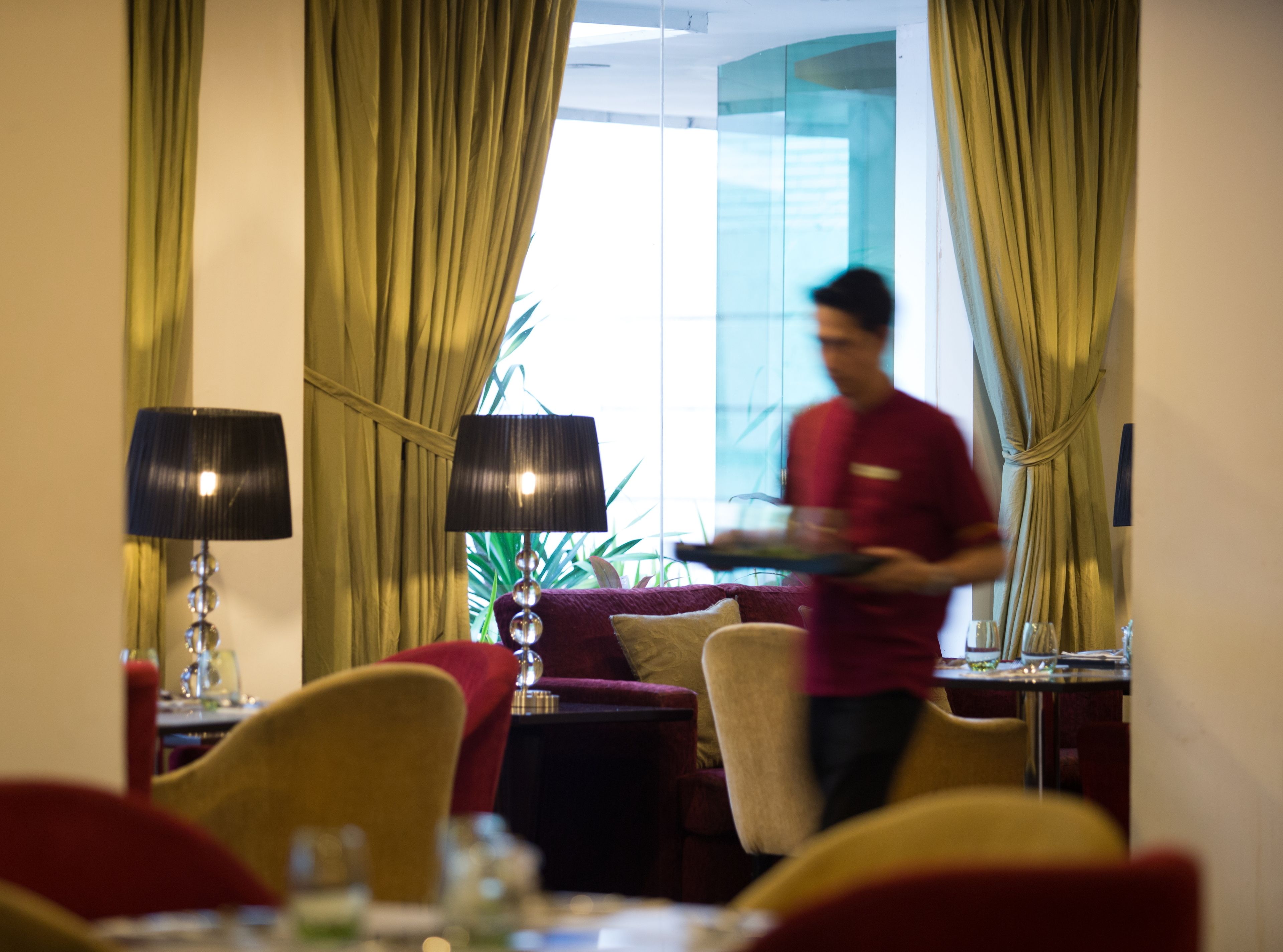 A hotel room with a dining area, a sofa, a lamp, and a large window with curtains.