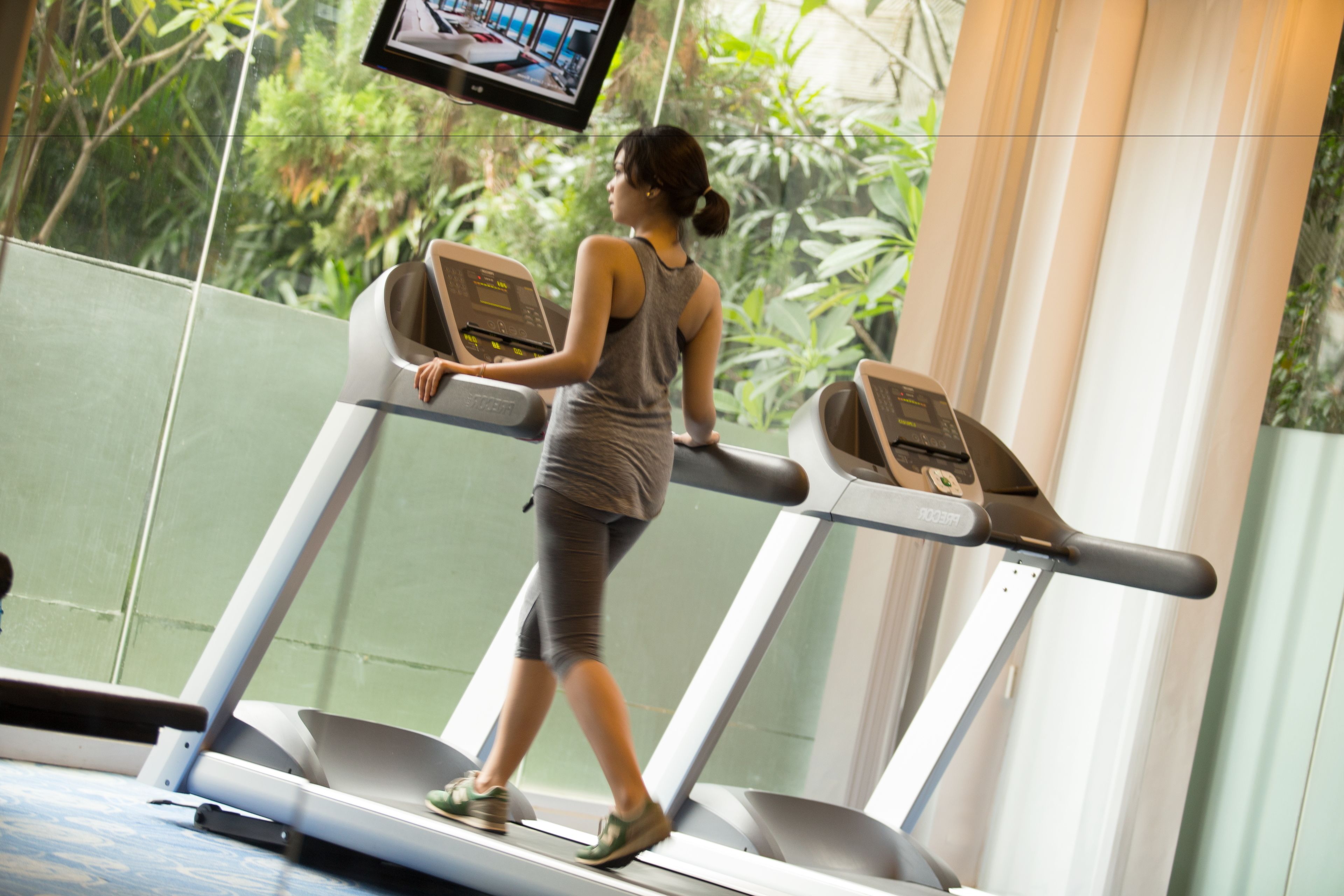 Eine Frau trainiert auf einem Laufband in einem Fitnessstudio mit großen Fenstern und Grünflächen im Hintergrund.