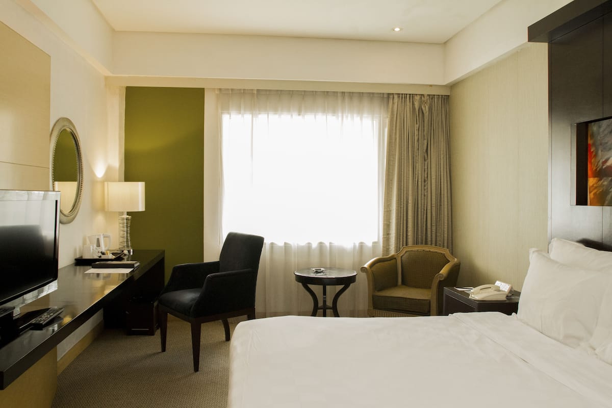 A hotel room with a bed, a TV, a desk with a lamp, two chairs, a small table, and a window with curtains.