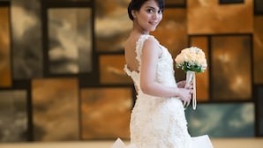 A person in a white wedding dress with a voluminous skirt, holding a bouquet, in a room with a modern, geometric wall design.