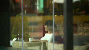 A person sitting by a window with a cup, a table with a glass, and a chair.