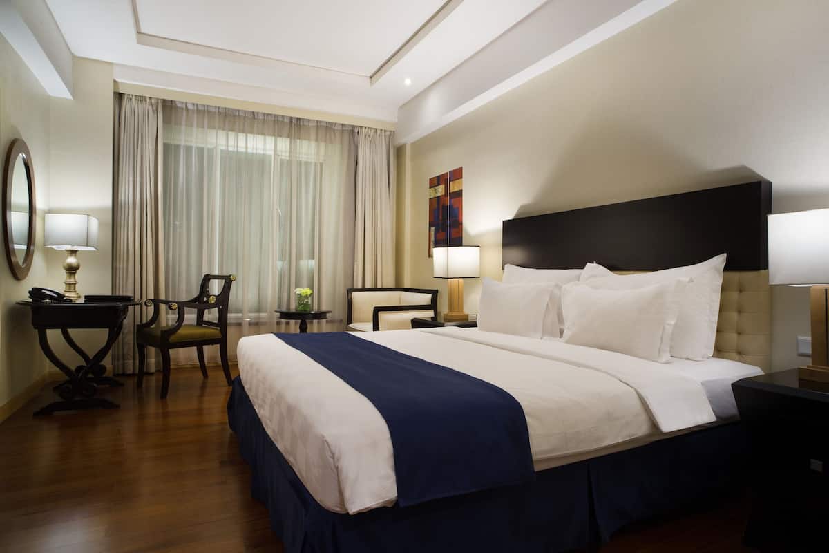 A hotel room with a large bed, a desk with a lamp, a chair, and a small table with a lamp.
