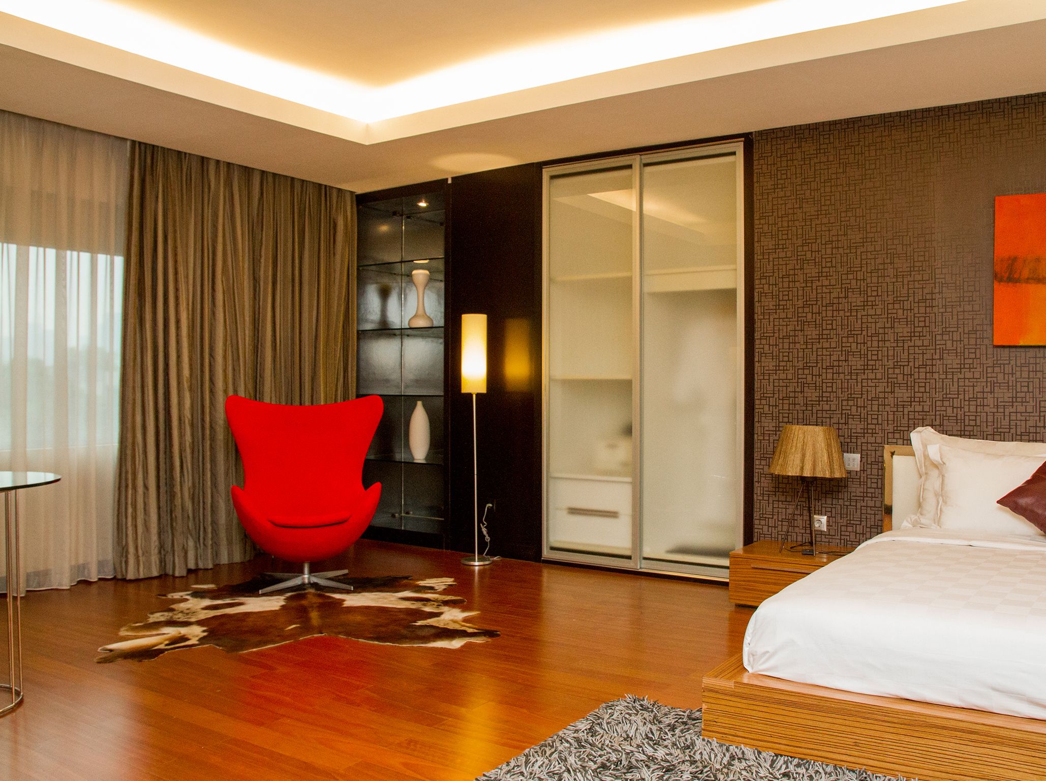 A modern hotel room with a large bed, a red chair, a wardrobe, and a glass door leading to a bathroom.
