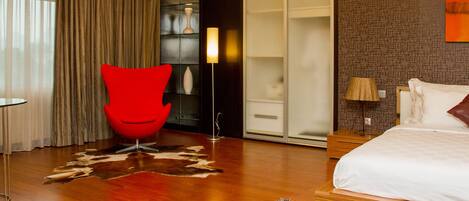 A modern hotel room with a large bed, a red chair, a wardrobe, and a glass door leading to a bathroom.