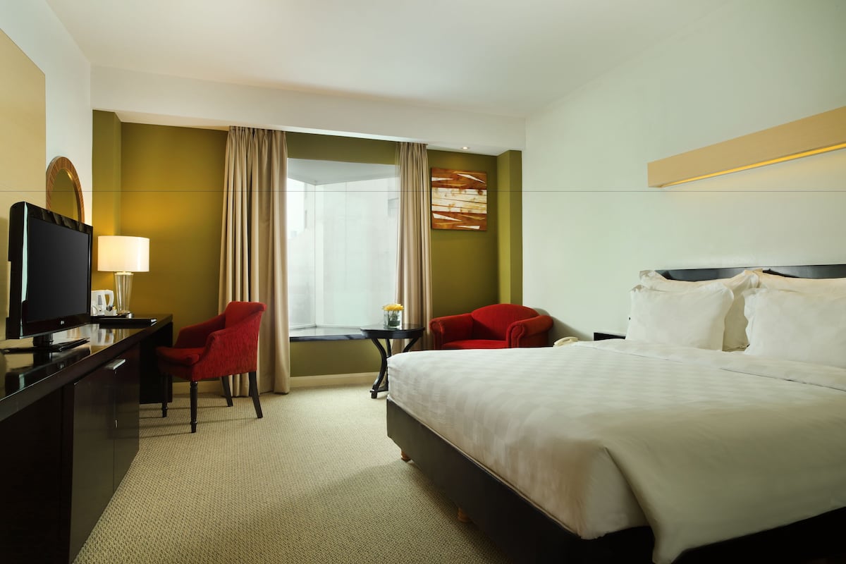 A hotel room with a bed, a desk with a television, two red chairs, a small table with a lamp, and a window with curtains.