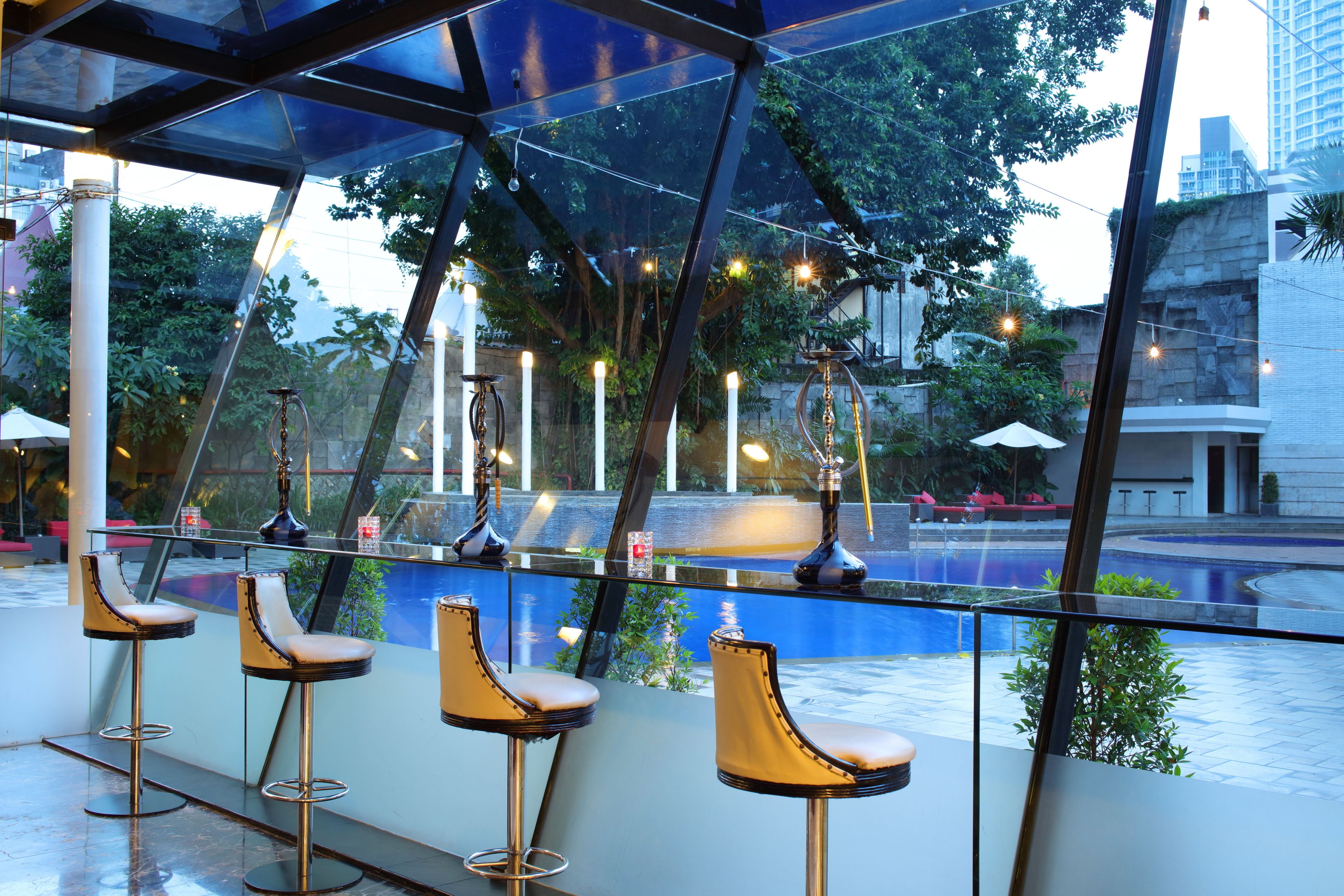 A modern bar area with a glass railing, multiple stools, and a view of a pool and greenery.