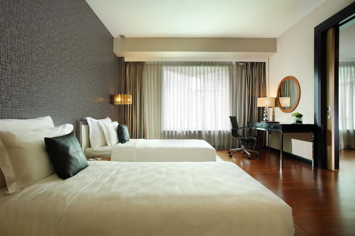 A hotel room with two beds, a desk, a chair, a lamp, and a large window with curtains.