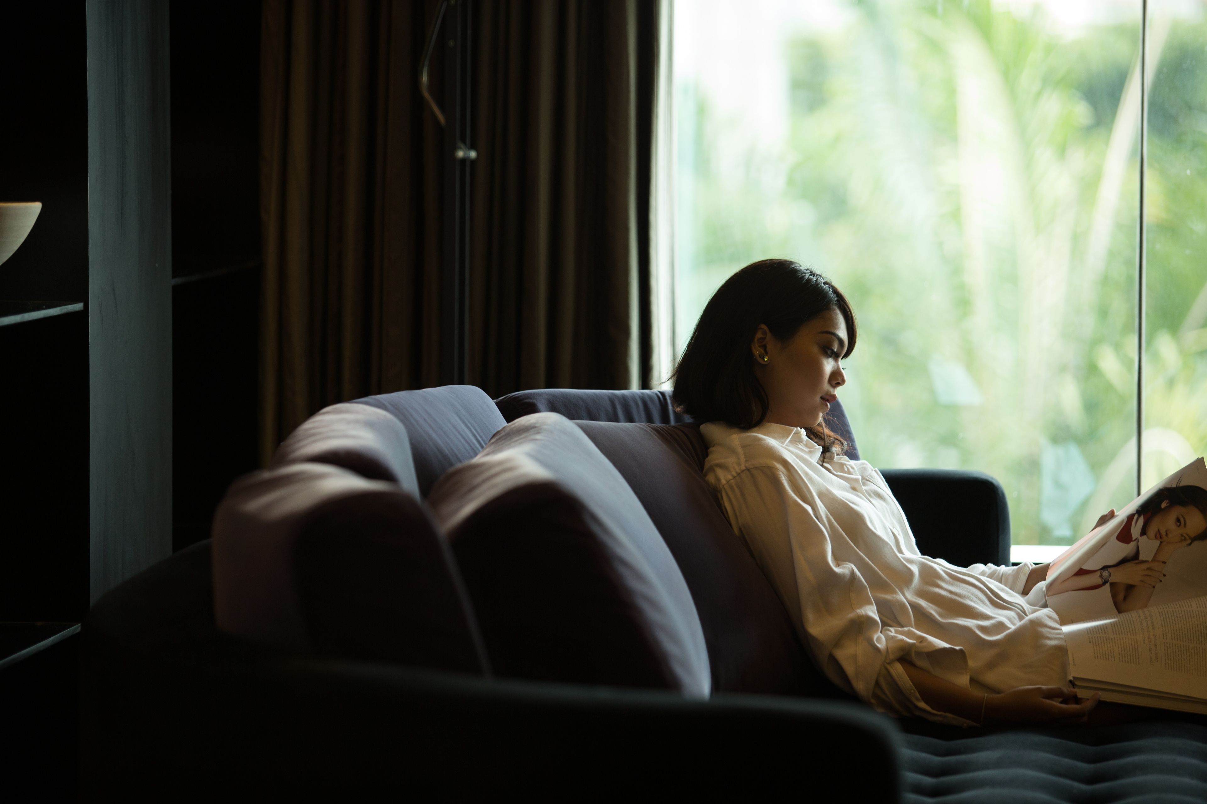 A person sitting on a couch in a room with a large window, reading a book.