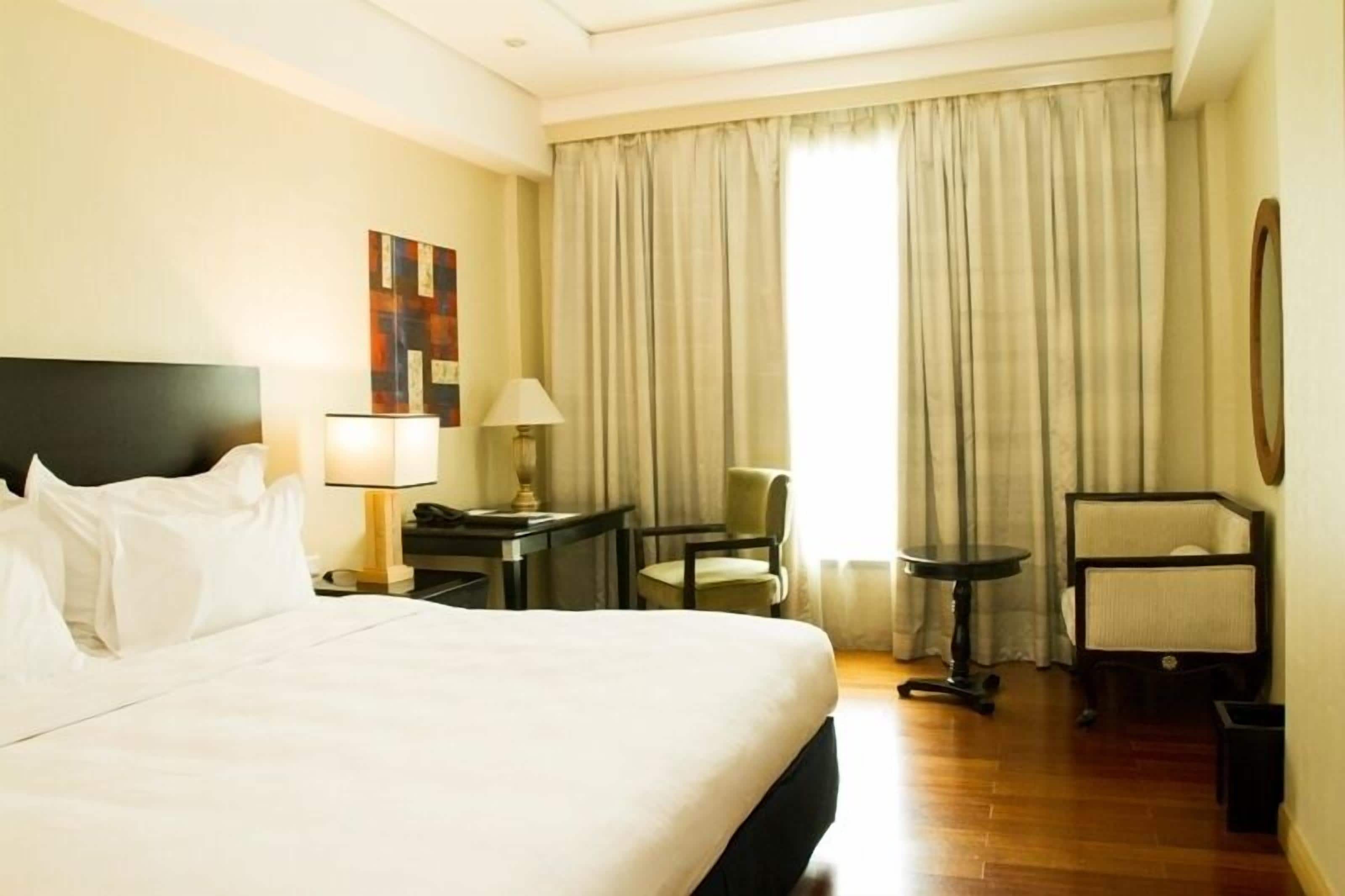 A hotel room with a large bed, a desk with a lamp, a chair, and a window with curtains.