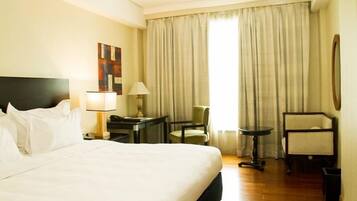 A hotel room with a large bed, a desk with a lamp, a chair, and a window with curtains.