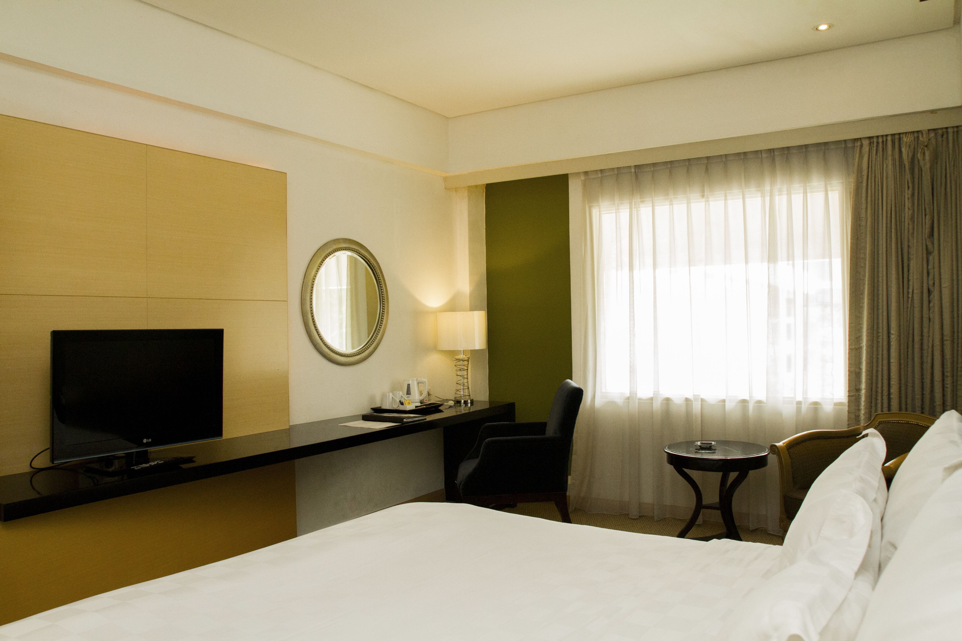 A hotel room with a bed, a desk with a TV, a chair, a small table, and a window with curtains.