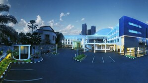 A modern hotel with a central glass facade, surrounded by palm trees and a parking lot.