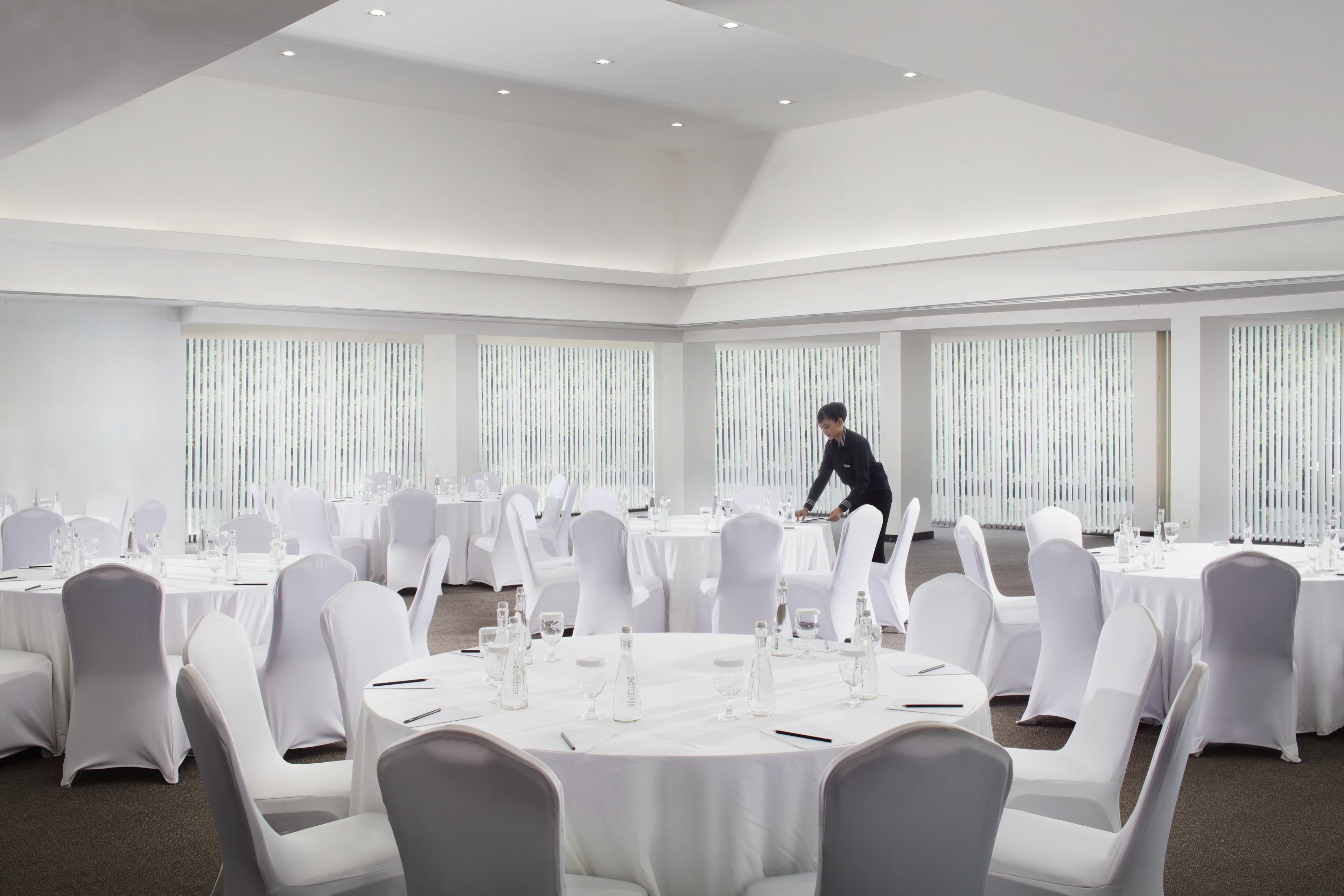 A large conference room with round tables, white chairs, and vertical blinds on the windows.