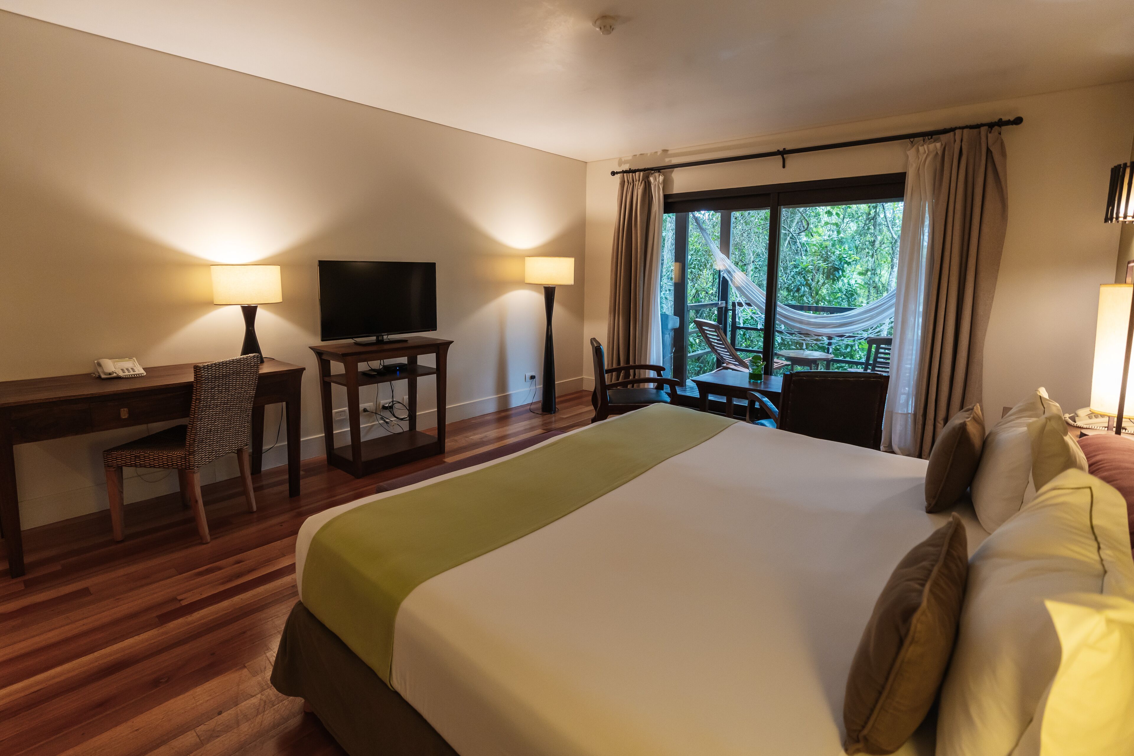 A hotel room with a large bed, a desk with a chair, a television, and a balcony with a hammock.