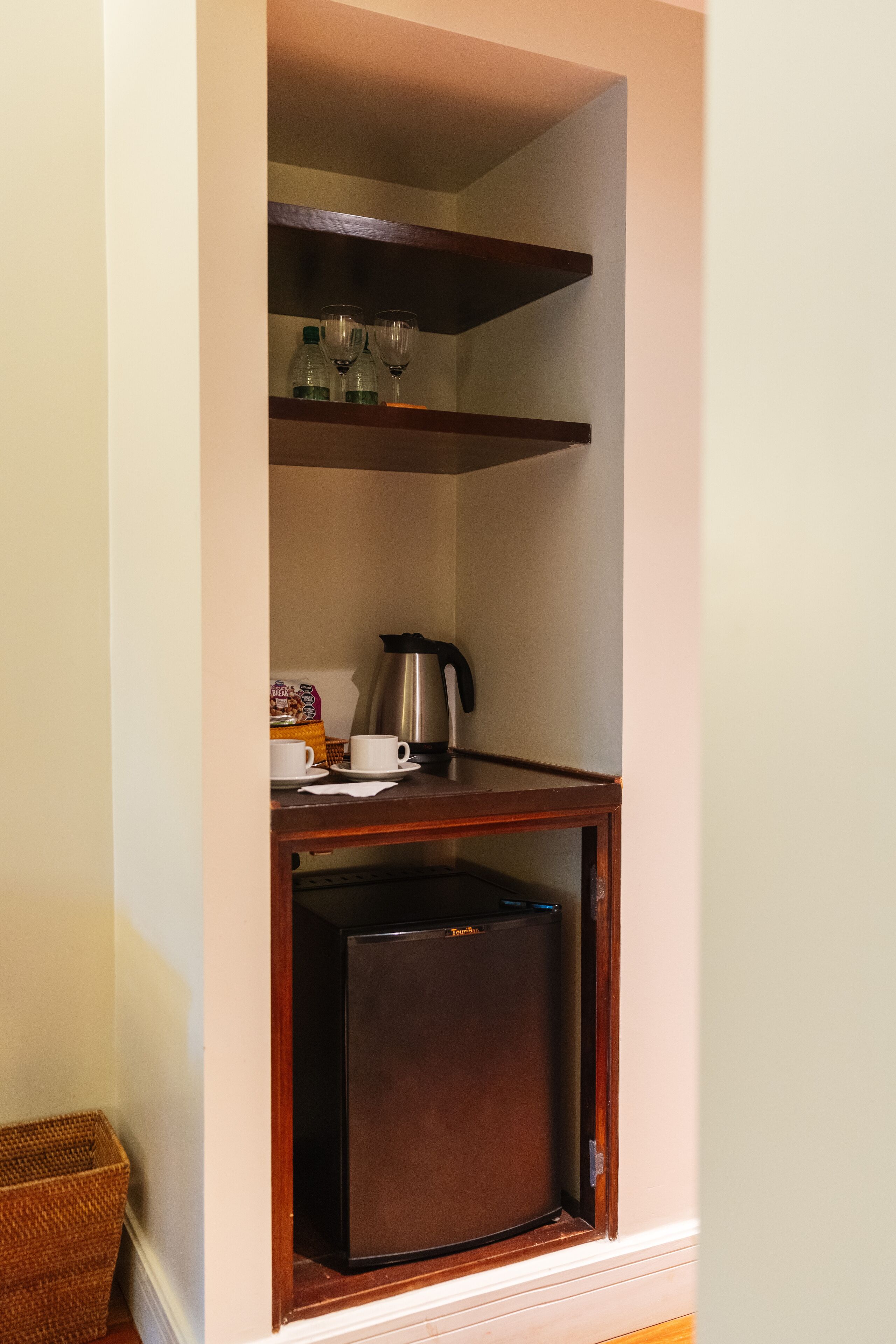 A compact kitchenette with a refrigerator, a coffee maker, and a shelf with glasses and bottles.