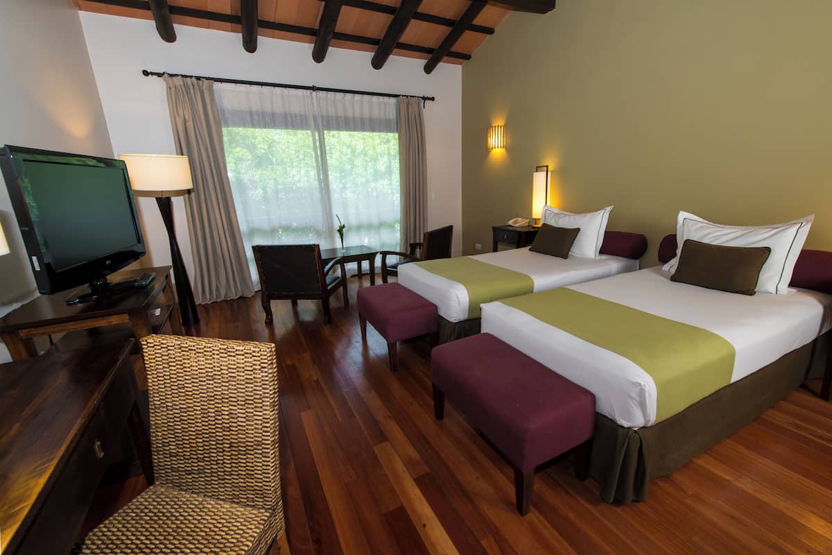 A hotel room with two beds, a desk with a television, a chair, and a window with curtains.