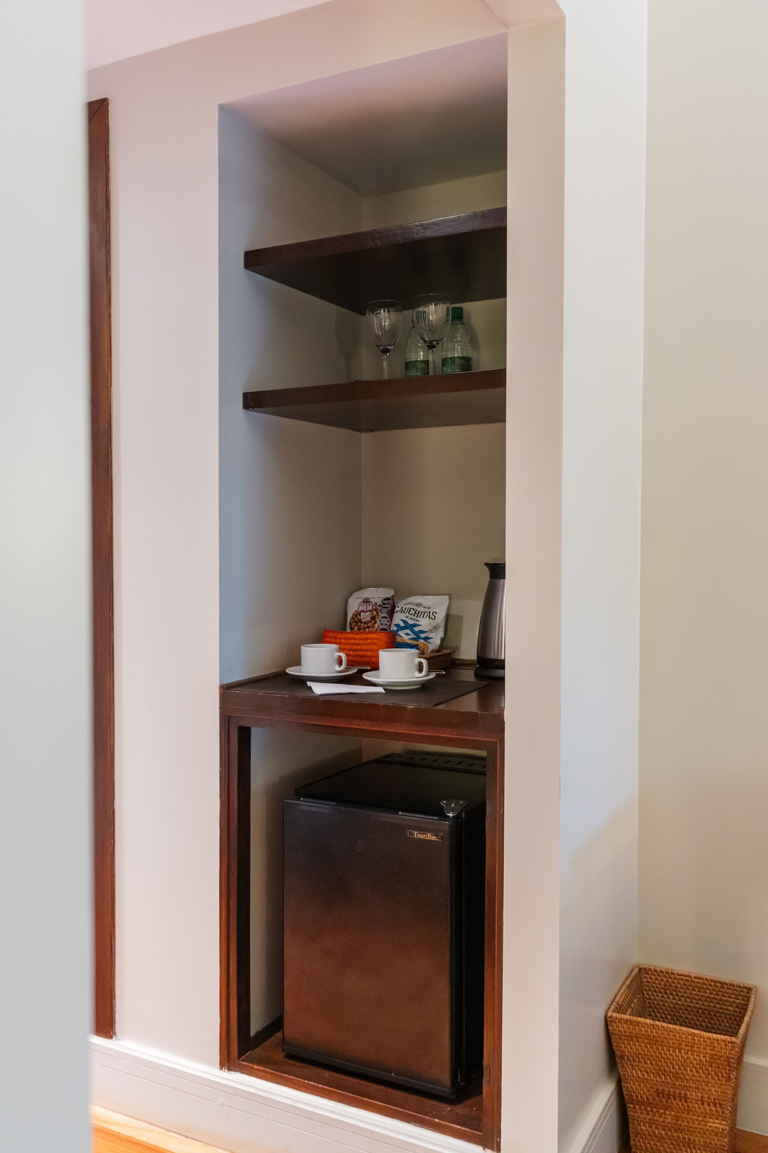 Una kitchenette compacta con un refrigerador integrado, una cafetera y una alacena con cristalería.