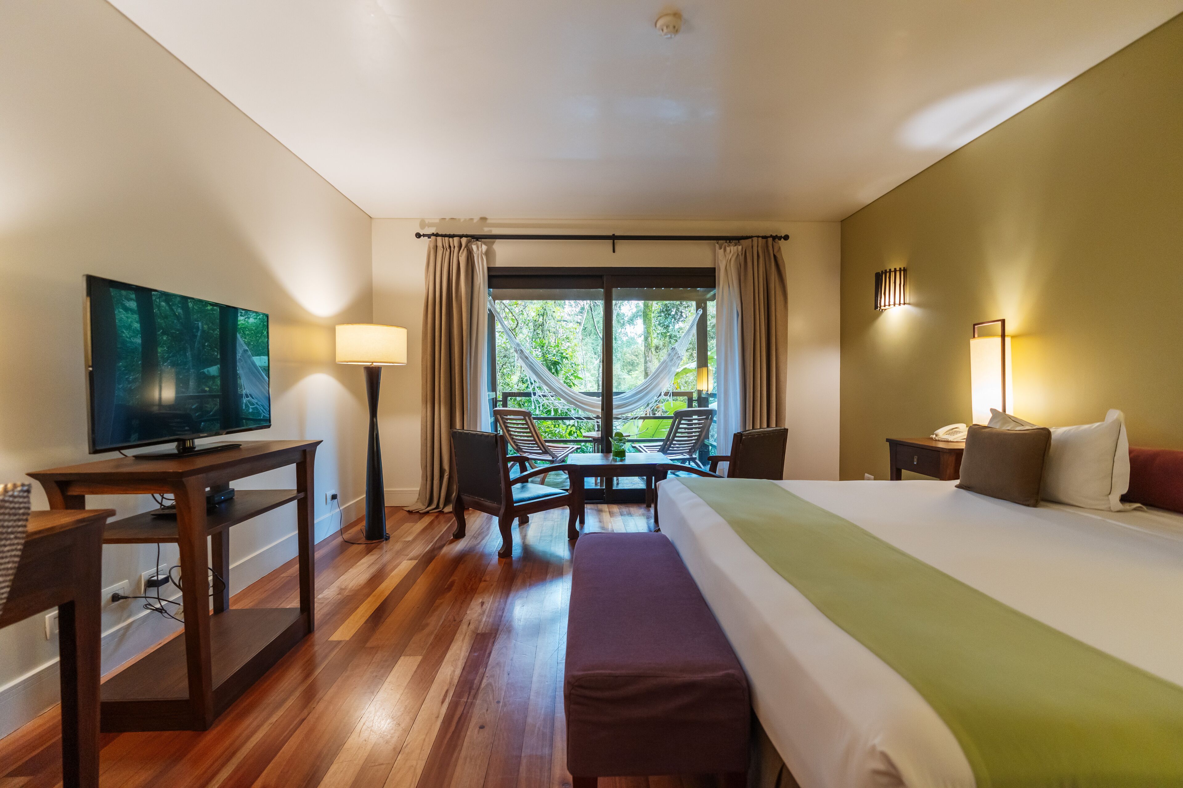 A hotel room with a bed, a desk with a television, a table with chairs, and a window with curtains.