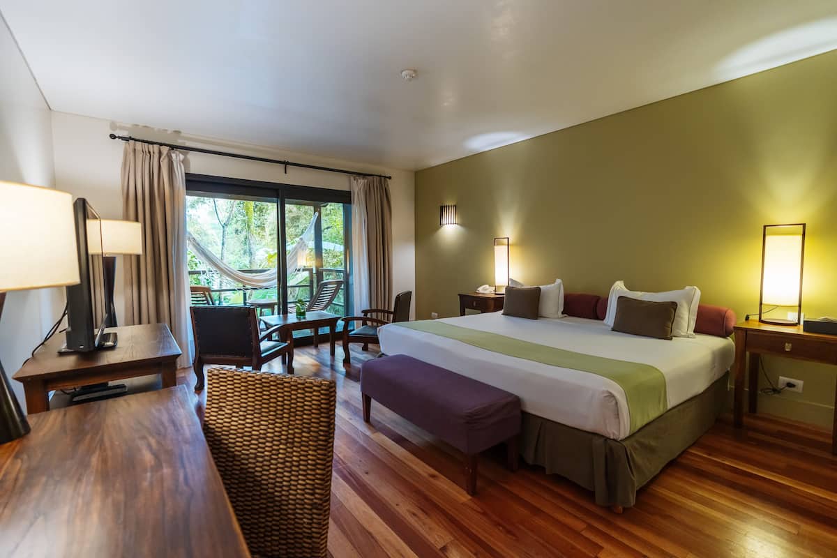 A hotel room with a large bed, a desk, a chair, and a balcony with a view of greenery.