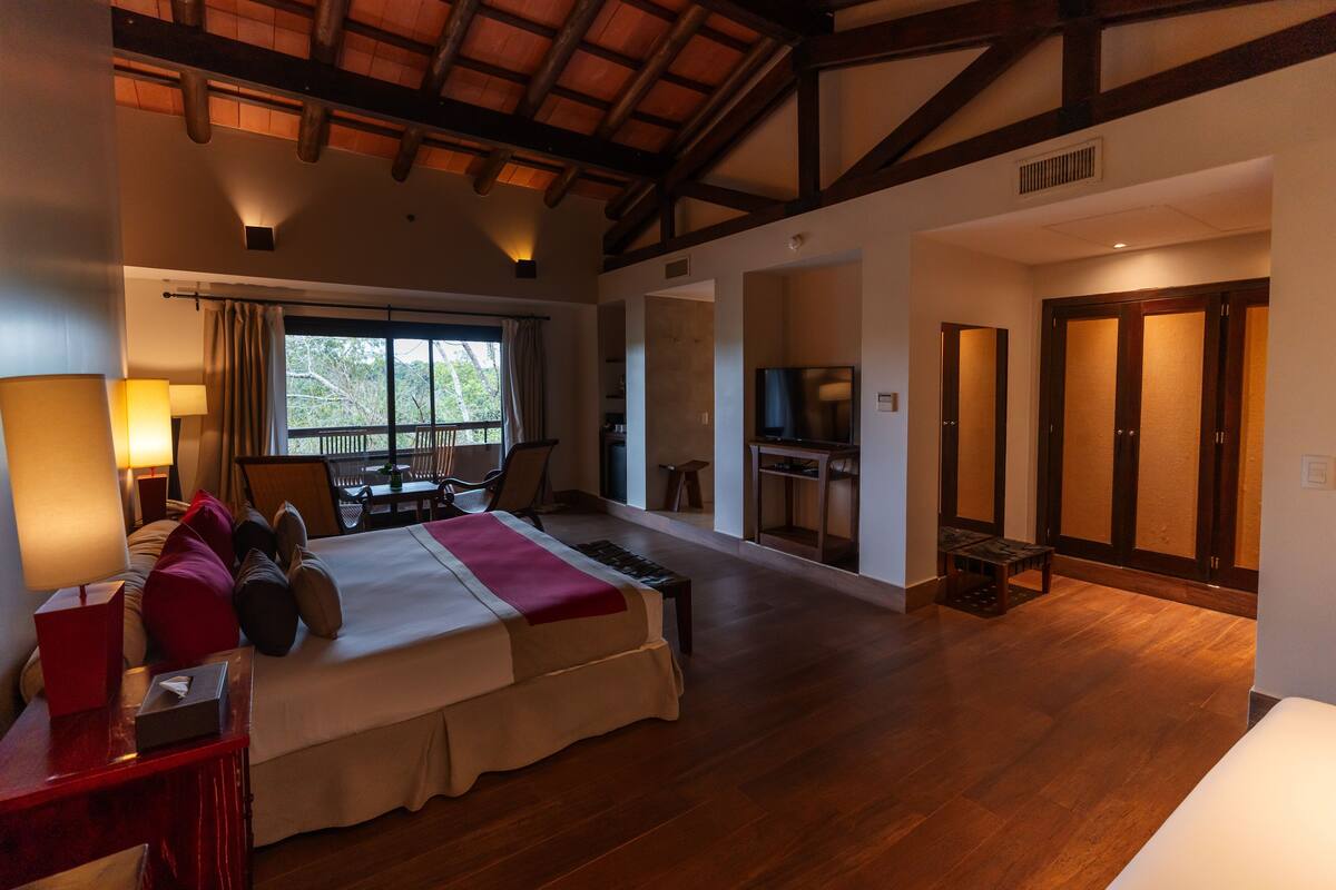 A spacious bedroom with a large bed, a desk, a TV, and a balcony with a view.
