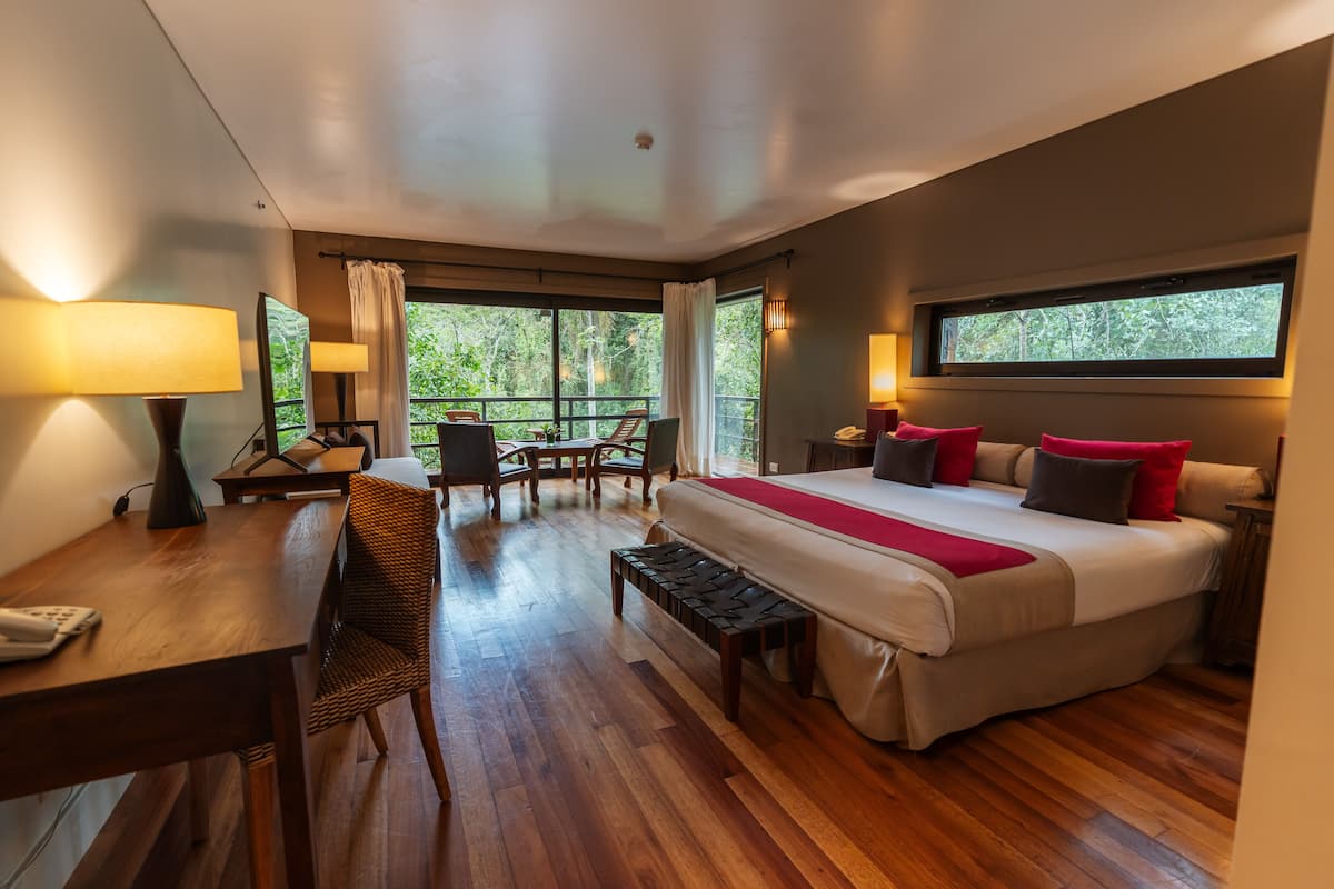 A bedroom with a large bed, a desk, a chair, and a balcony with a view of trees.