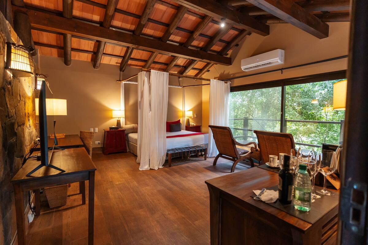 A cozy room with a wooden ceiling, a bed with white curtains, a dining table with chairs, and a view of greenery outside.
