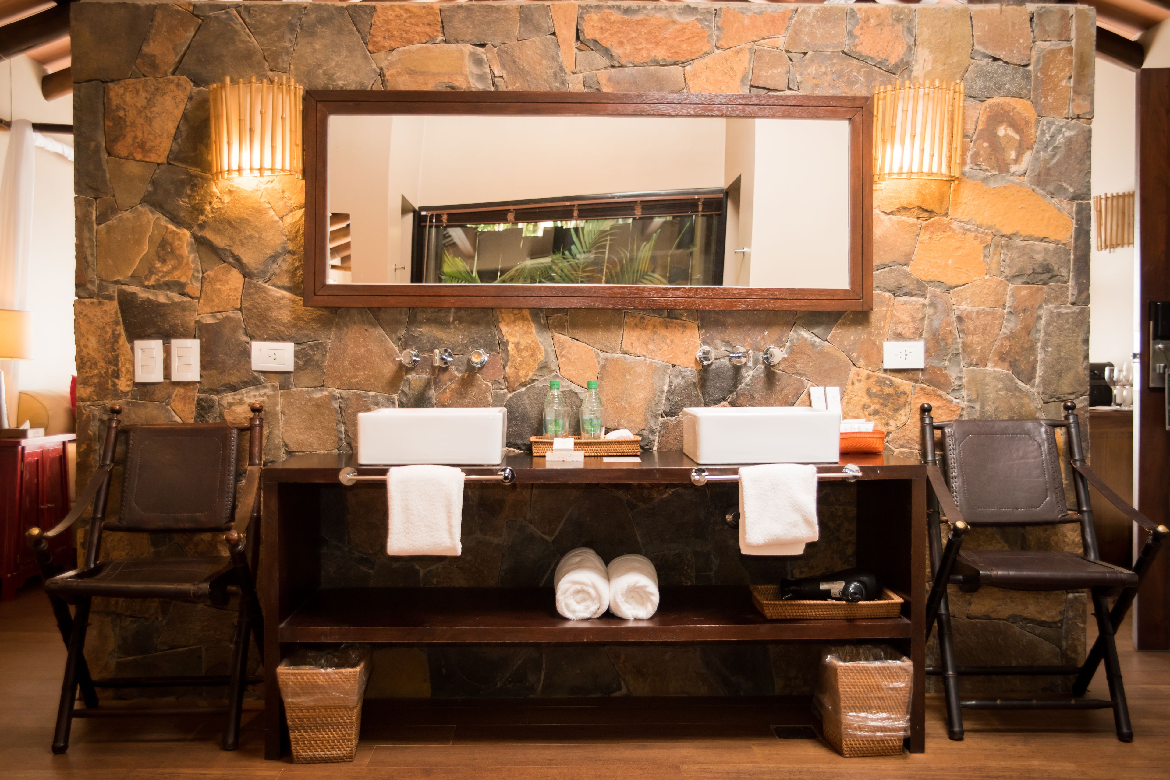 A stone wall with a mirror, two sinks, and two chairs.