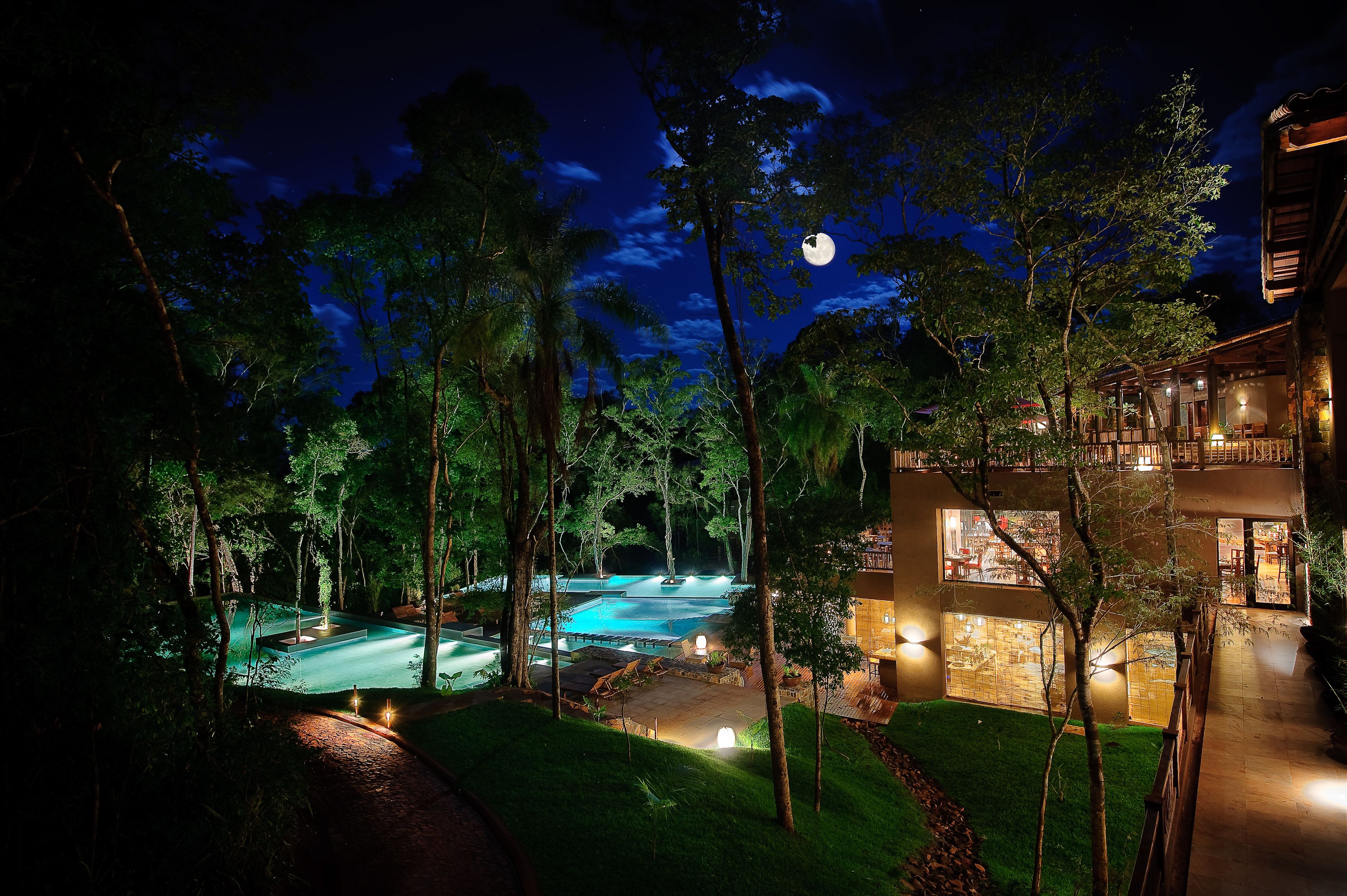 A nighttime view of a resort with a swimming pool, surrounded by trees and a building with multiple floors and balconies.