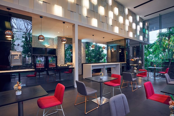 A modern restaurant with a bar area, red and grey chairs, and a view of greenery through large windows.