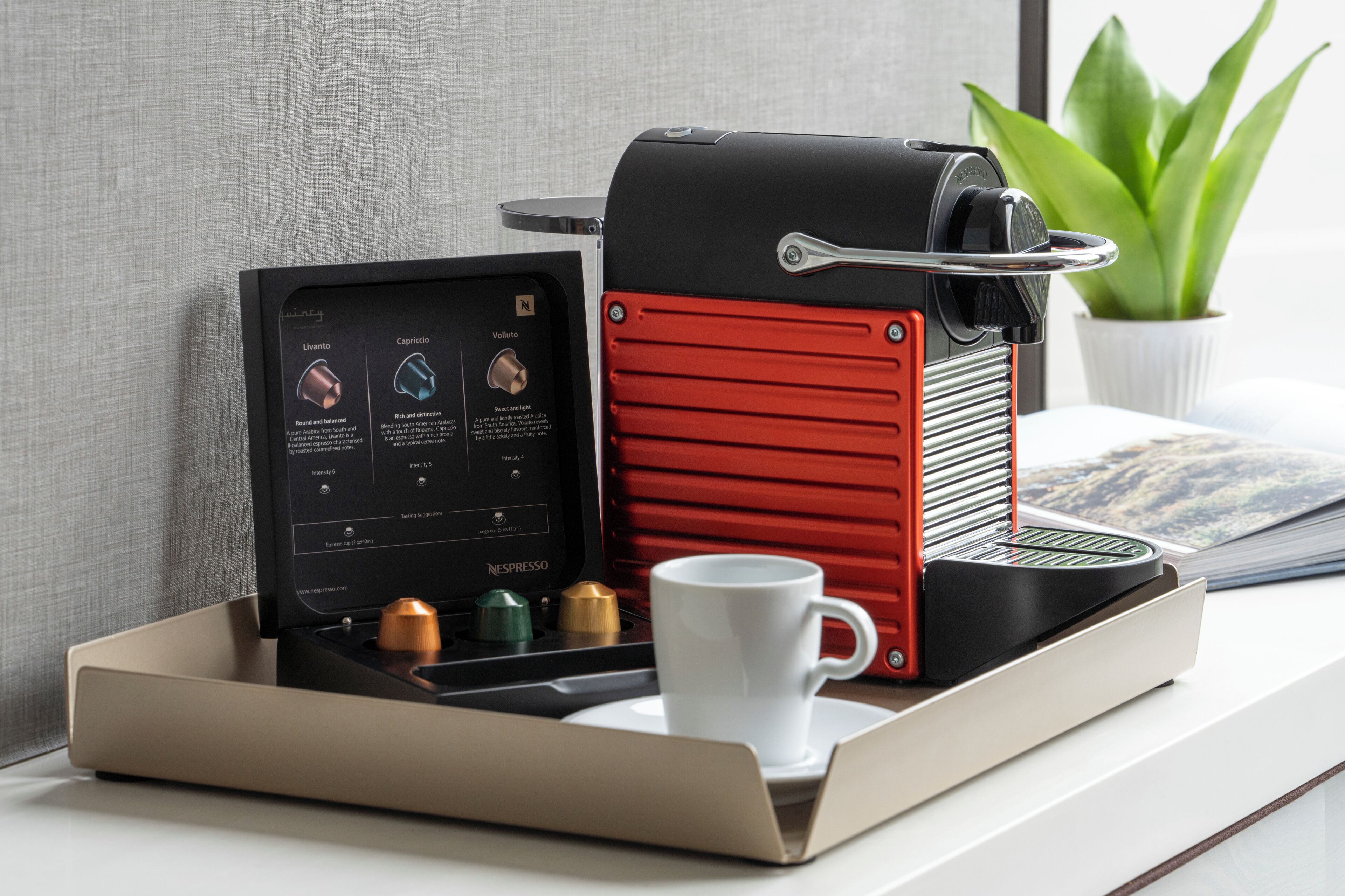 Una máquina de café Nespresso de diseño rojo y negro, una taza blanca y una bandeja con una planta y una revista.