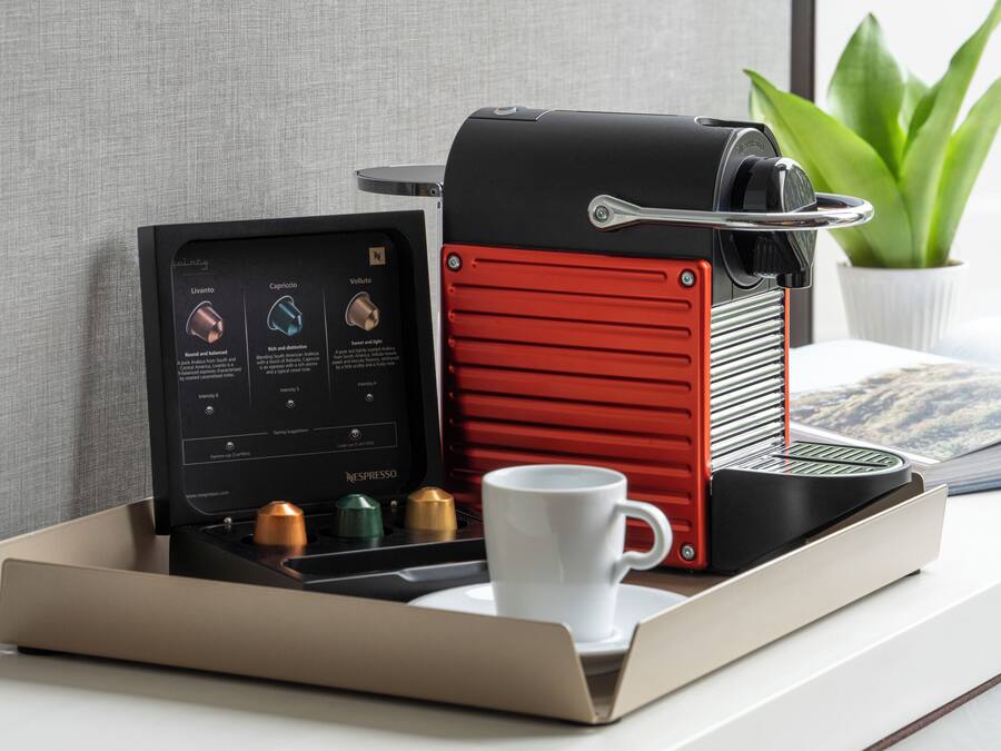 Una máquina de café Nespresso de diseño rojo y negro, una taza blanca y una bandeja con una planta y una revista.