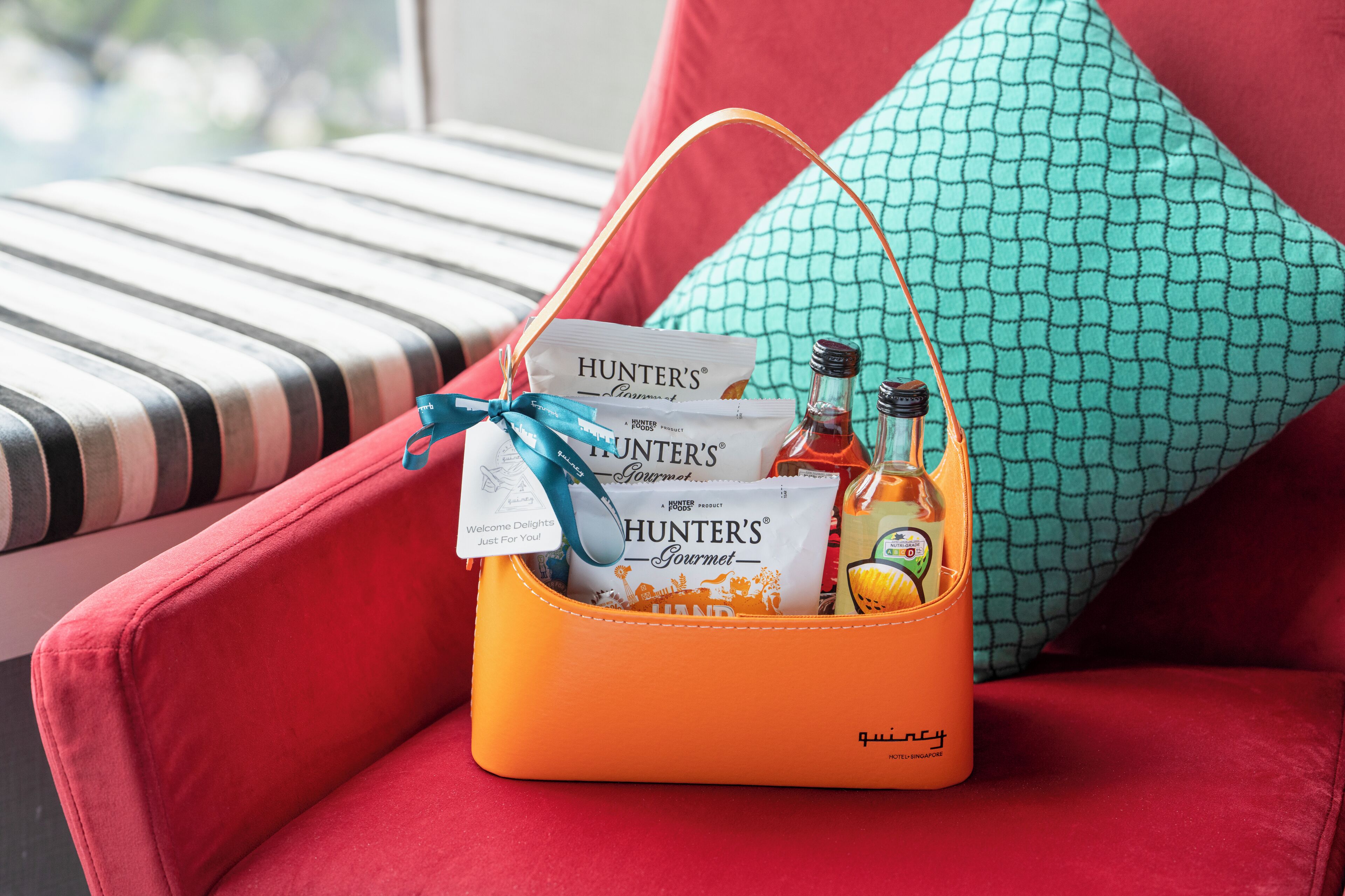 Een rode stoel met een gestreept kussen, een teal kussen met een patroon, en een feloranje tas met daarin een fles, een fles en een kaartje met de tekst 'Hunter's Gourmet'.
