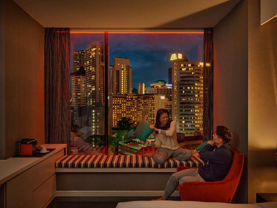 A modern hotel room with a city view, a built-in bench, a red armchair, and a small table with a cup and a book.