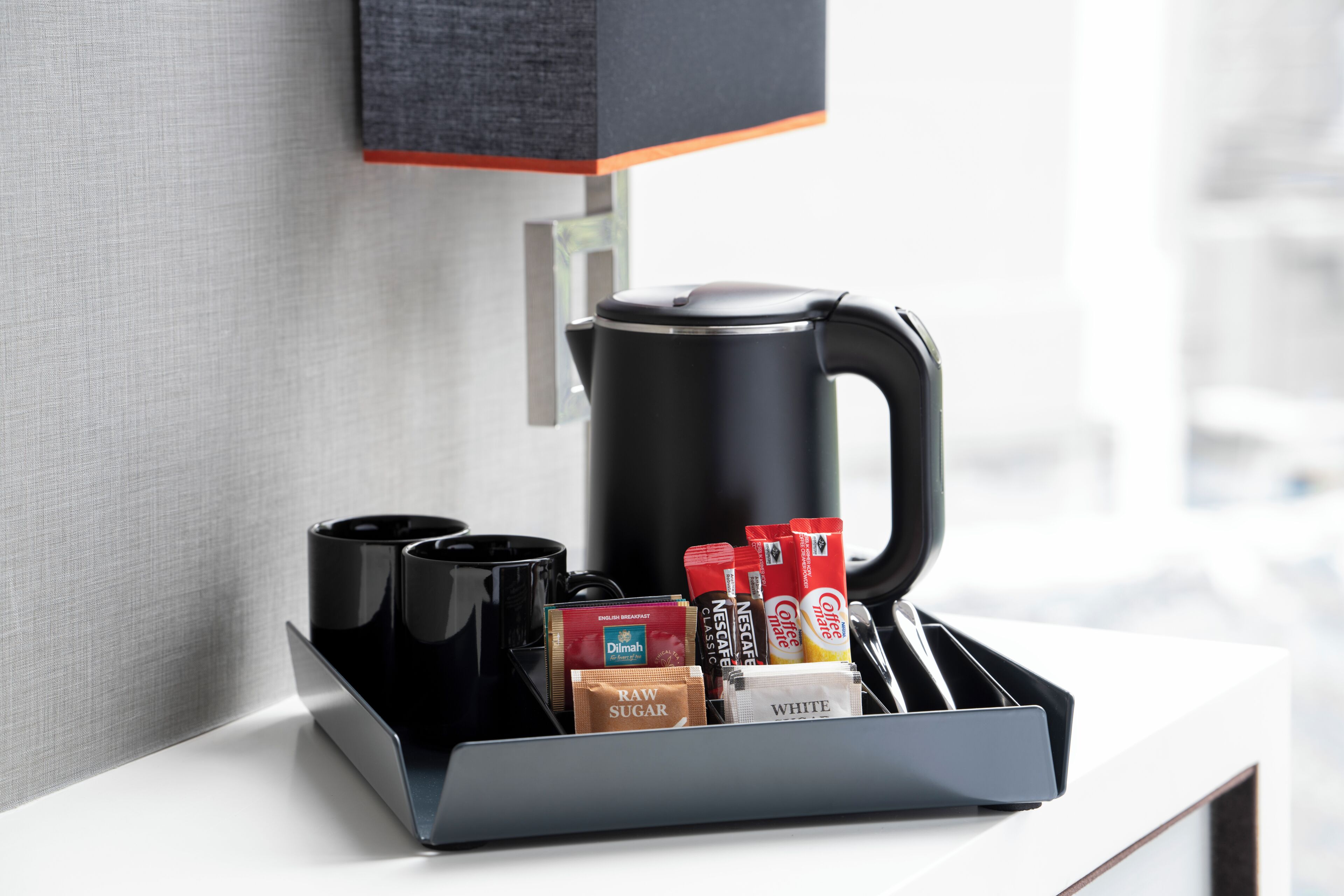Una estación de café con una cafetera negra, dos tazas negras, y variedad de cápsulas de café y sobres de azúcar.