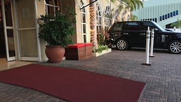 A hotel entrance with a red carpet, a large potted plant, and a black SUV parked outside.