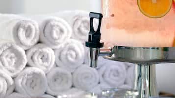 A beverage dispenser with a clear glass filled with a pinkish drink, garnished with orange slices, and a black handle. In the background, there are neatly rolled white towels.