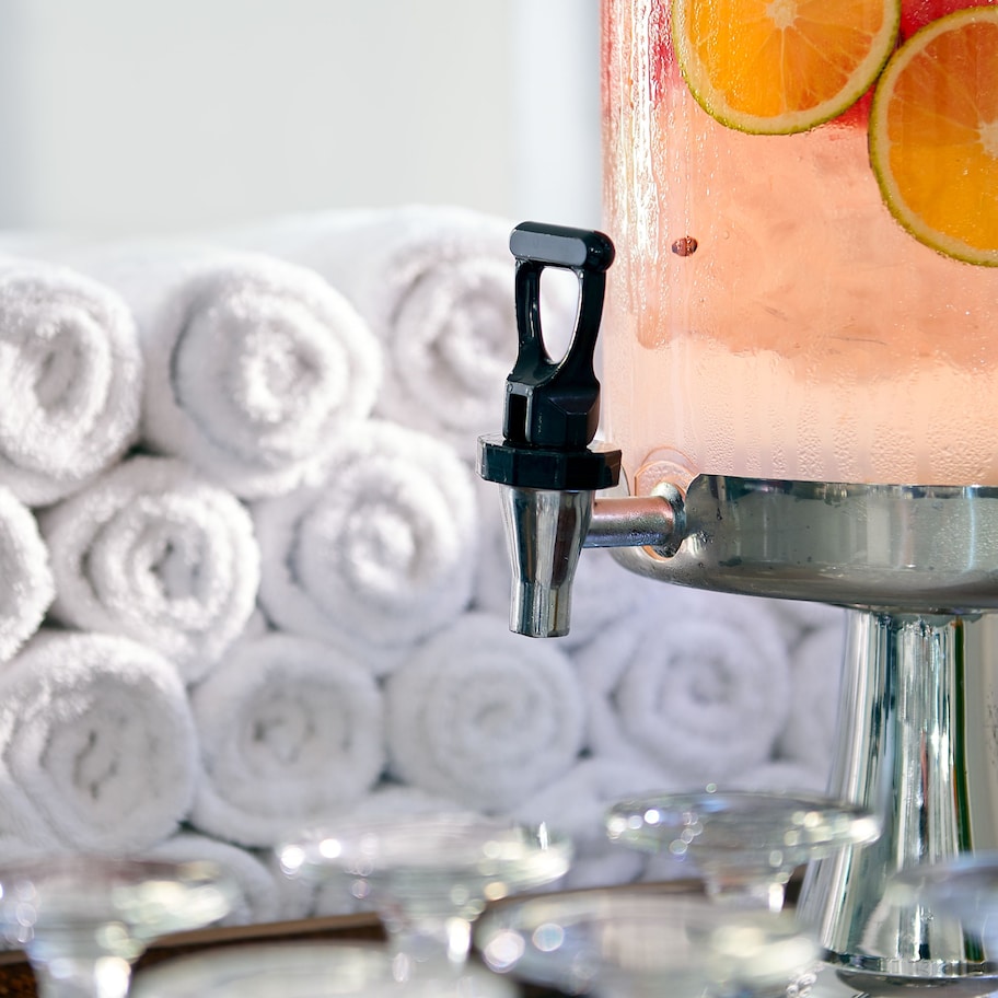 A beverage dispenser with a clear glass filled with a pinkish drink, garnished with orange slices, and a black handle. In the background, there are neatly rolled white towels.