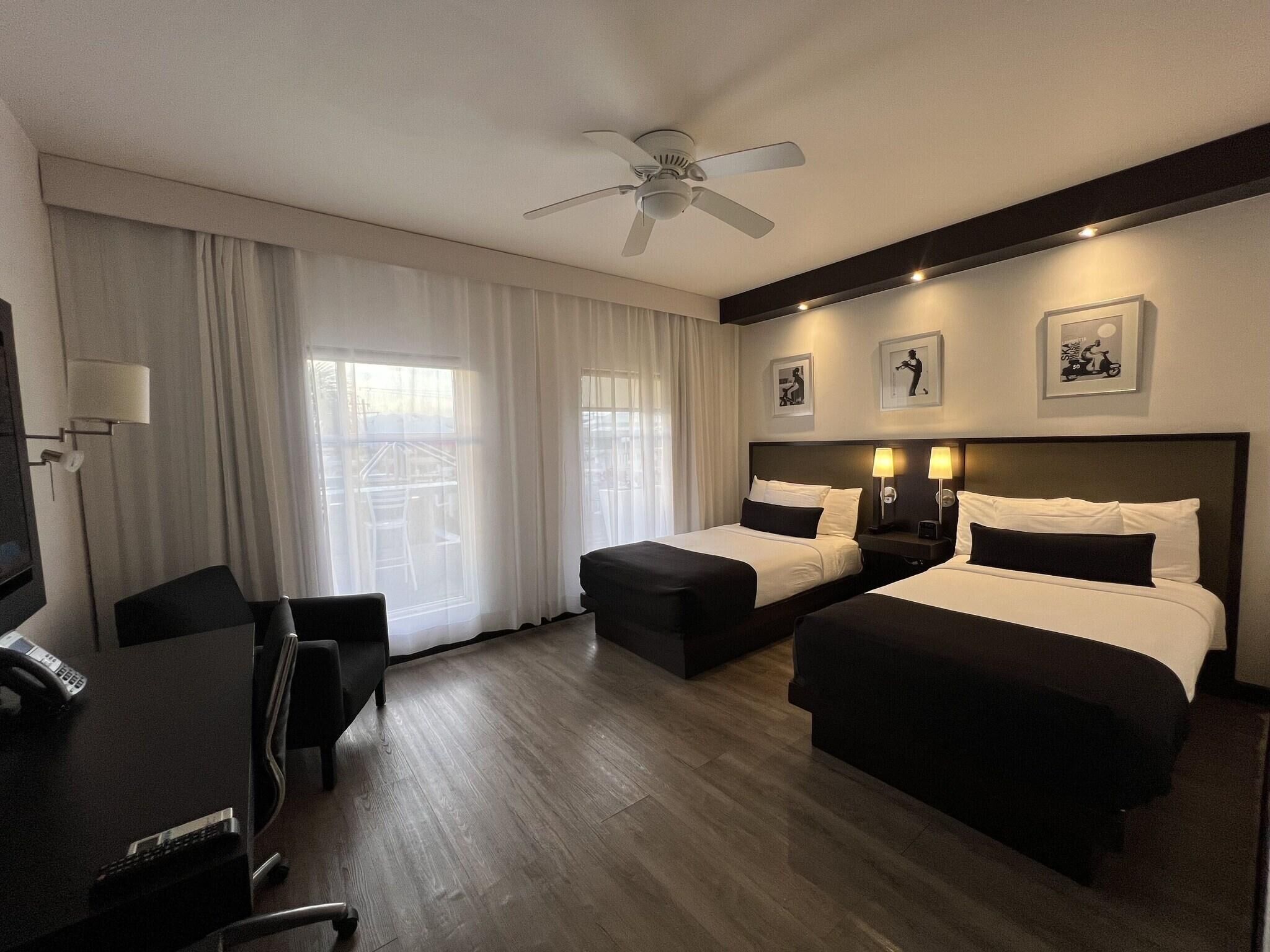 A hotel room with two beds, a desk with a telephone, a chair, a ceiling fan, and large windows with curtains.