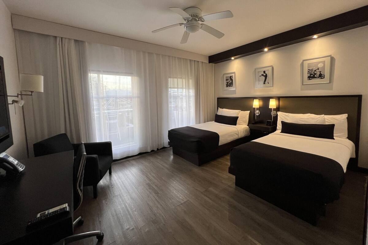 A hotel room with two beds, a desk with a telephone, a chair, a ceiling fan, and large windows with curtains.